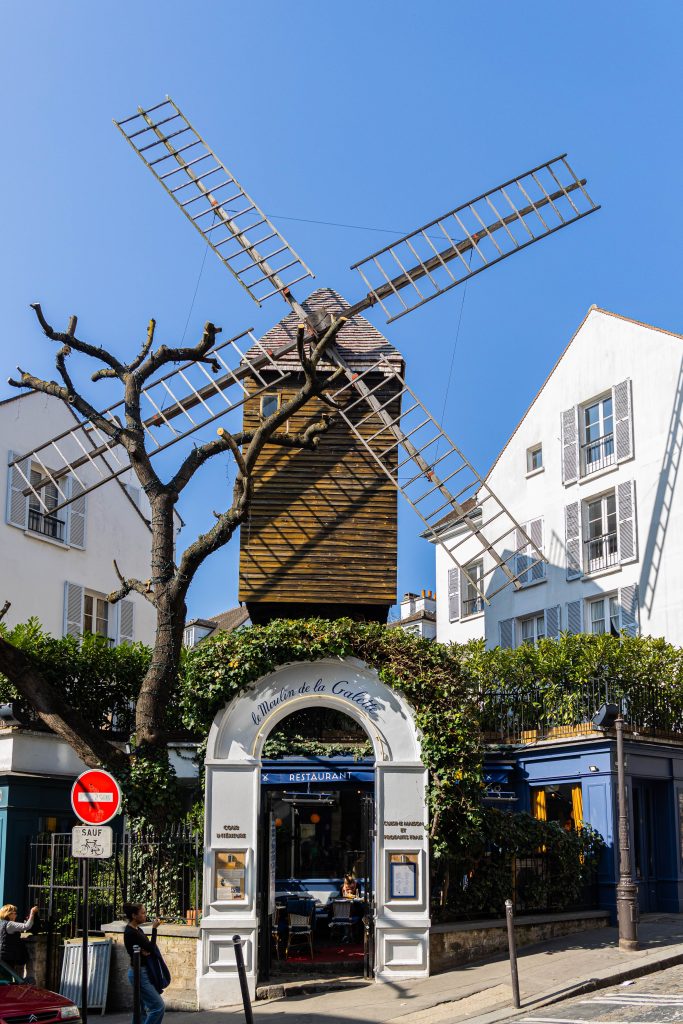 Image de face devant le restaurant du Moulin de la Galette montrant l'entrée dans la partie basse de l'image et le Moulin dans la partie haute.